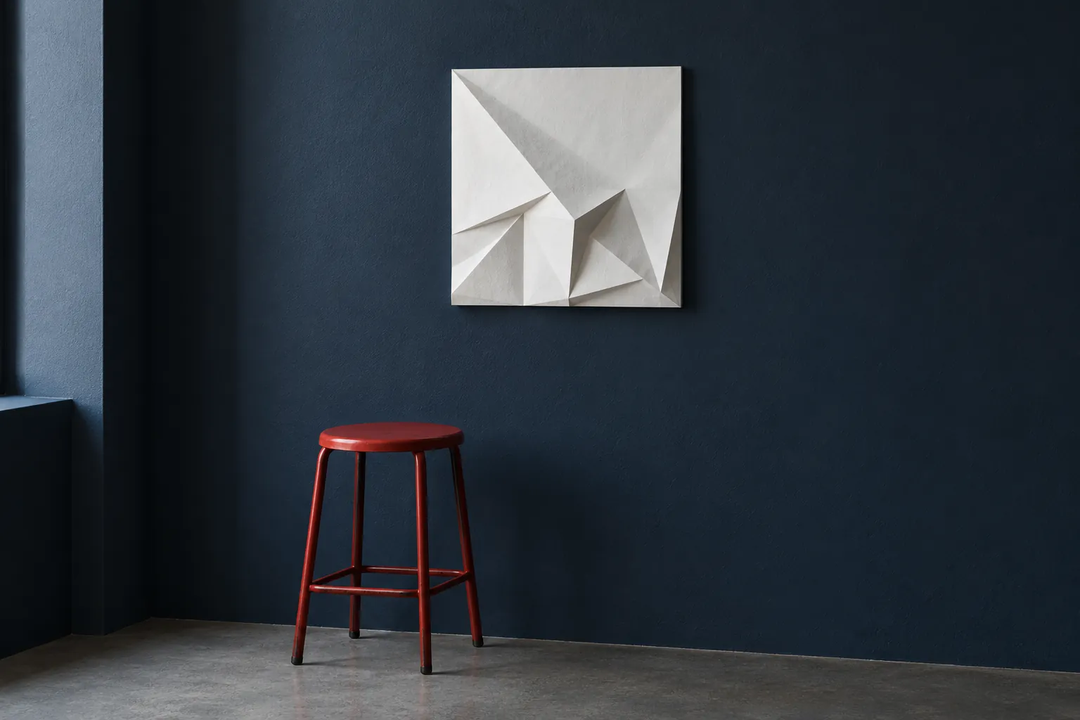 Illustration: A small original white folded-paper relief hangs on a navy gallery wall above a lone red steel stool.