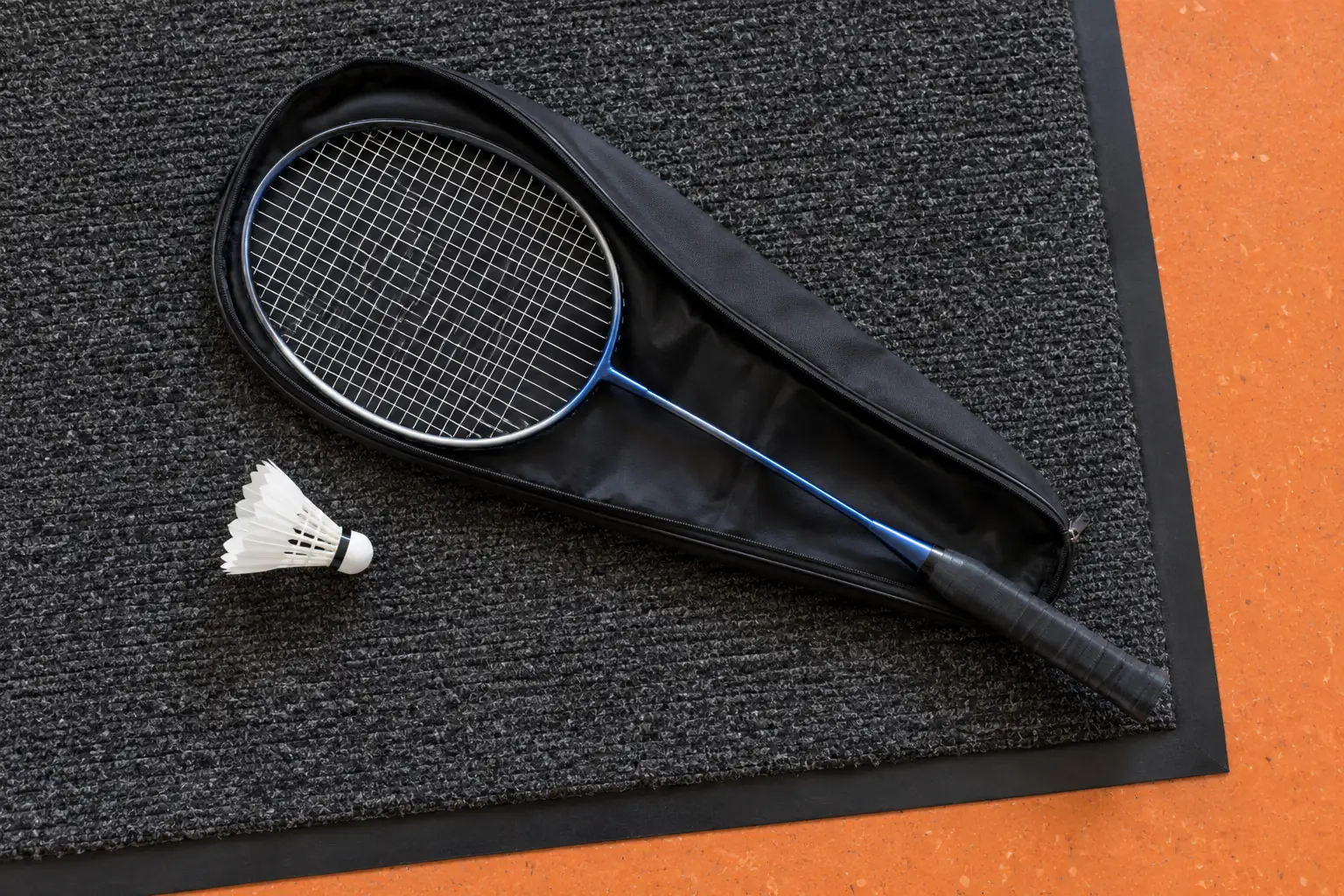 Illustration: A plain slim oval badminton racket in a half-open sleeve lies on a charcoal entrance mat beside a white shuttlecock.