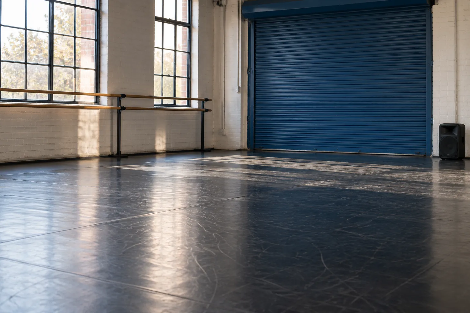 Illustration: An empty street-dance rehearsal space has a dark sprung floor, a blue roller door and a compact unbranded speaker at the perimeter, photographed diagonally.