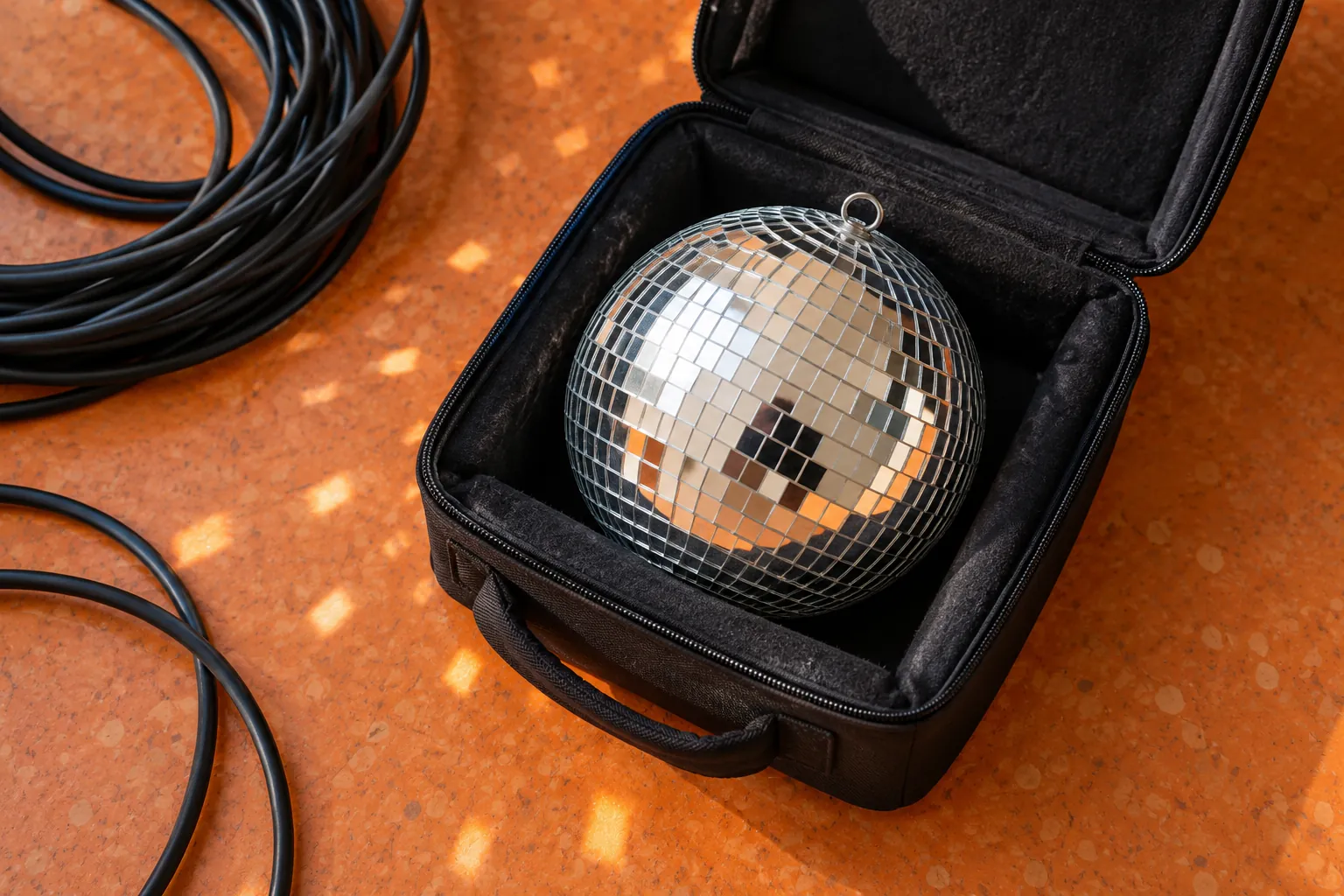 Illustration: A small mirrored disco ball rests safely in an open black padded case on orange linoleum beside coiled cables, photographed close overhead with no people reflected.