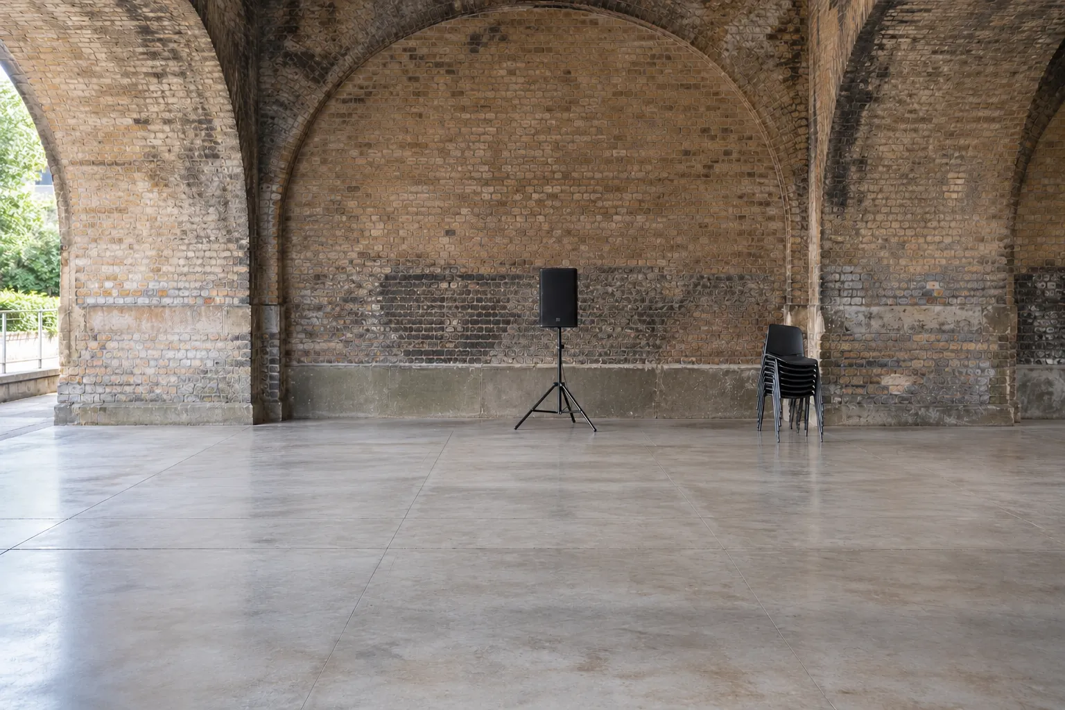 Illustration: An open concrete practice floor beneath brick arches, with a speaker and stacked chairs stored at the back.