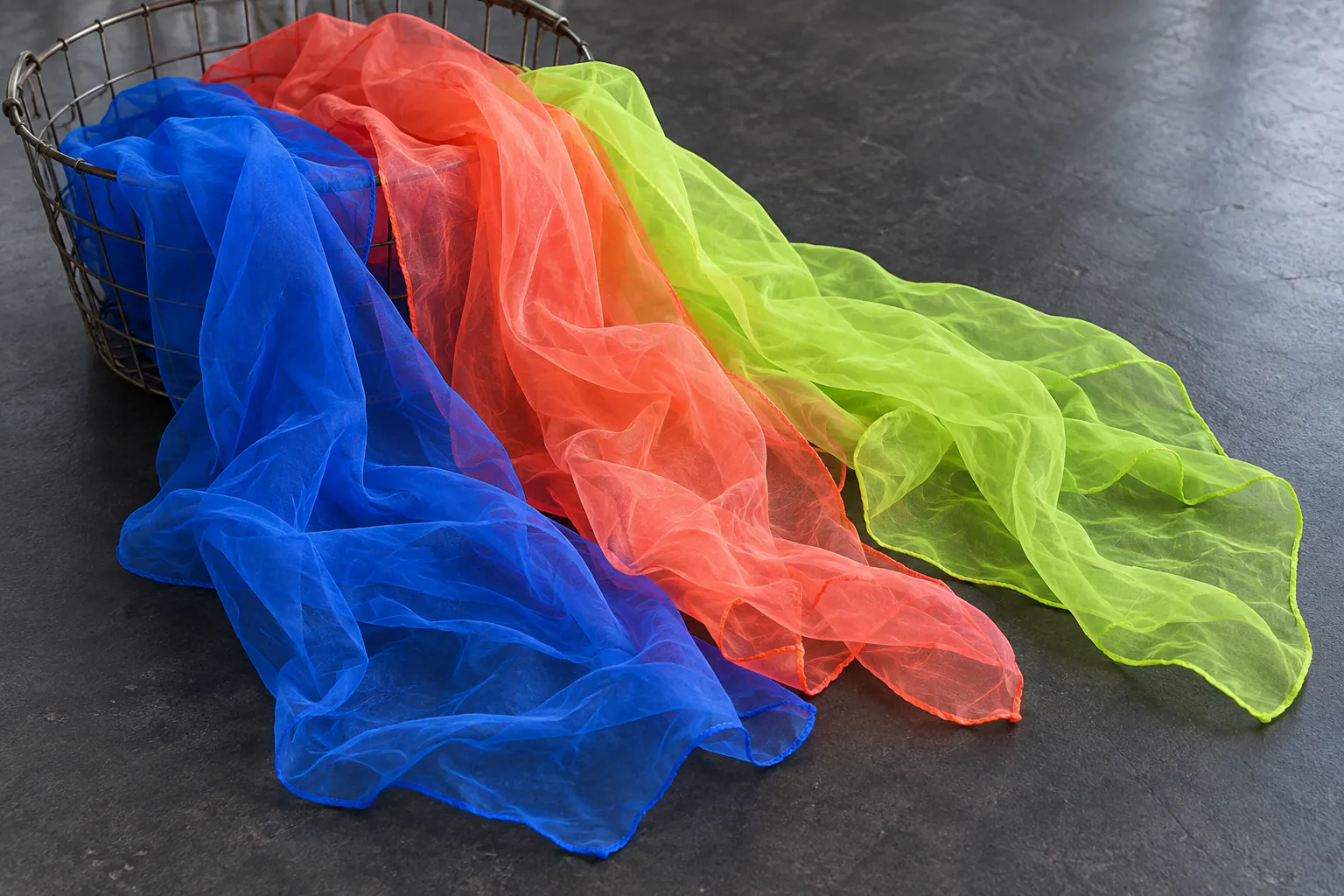 Illustration: Bright blue, coral and lime movement scarves spill gently from a wire basket onto a dark studio floor, photographed close overhead with no unrelated exercise props.