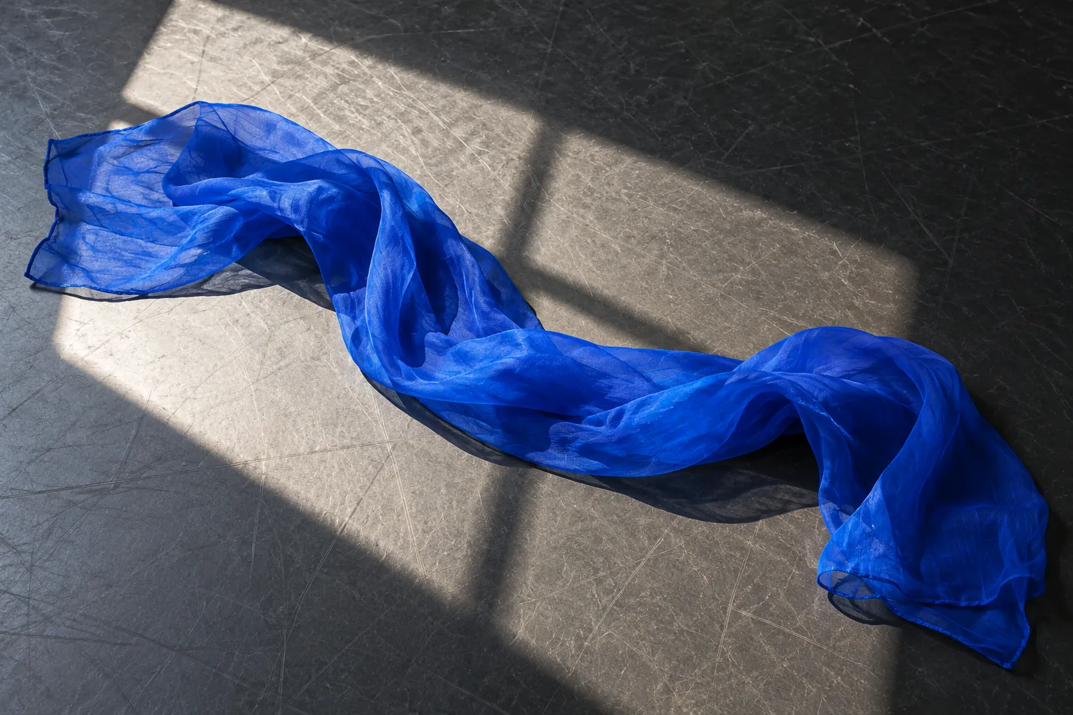 Illustration: A single cobalt movement scarf lies in a loose curve across a charcoal dance floor cut by a rectangular patch of sunlight, photographed overhead as an activity detail.