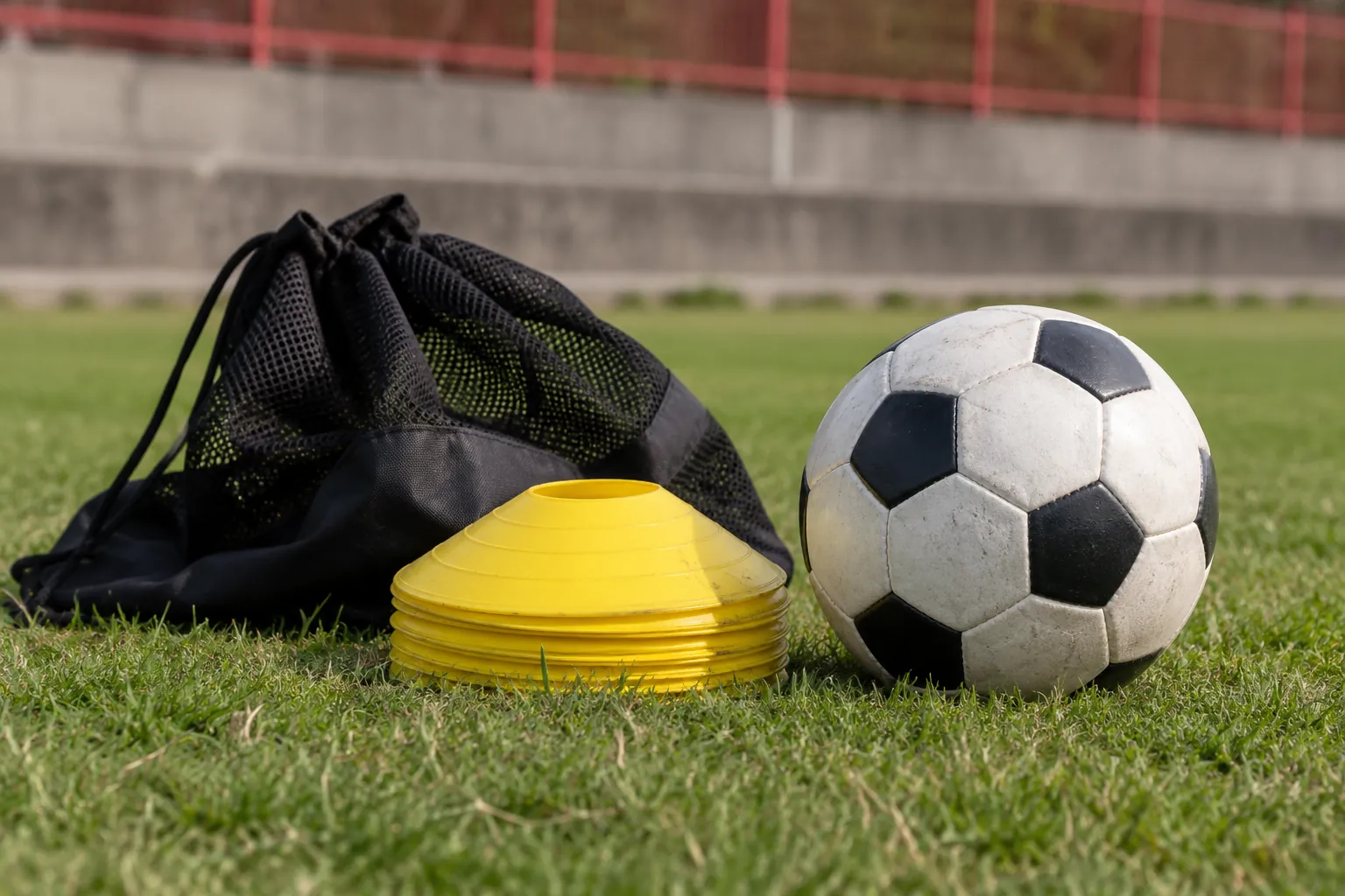 Illustration: A black-and-white football and a short stack of flat yellow field markers rest beside a dark mesh bag on closely cut grass.