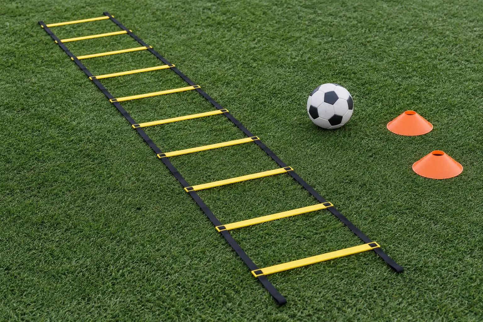 Illustration: A flat yellow football agility ladder lies straight on green turf beside a plain football and two low orange cones.