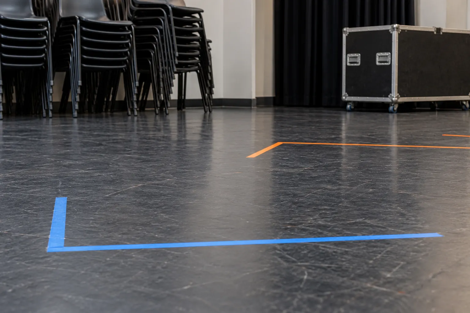 Illustration: Blue and orange tape forms simple stage-position marks on a charcoal floor beside stacked plastic chairs and a closed prop crate.