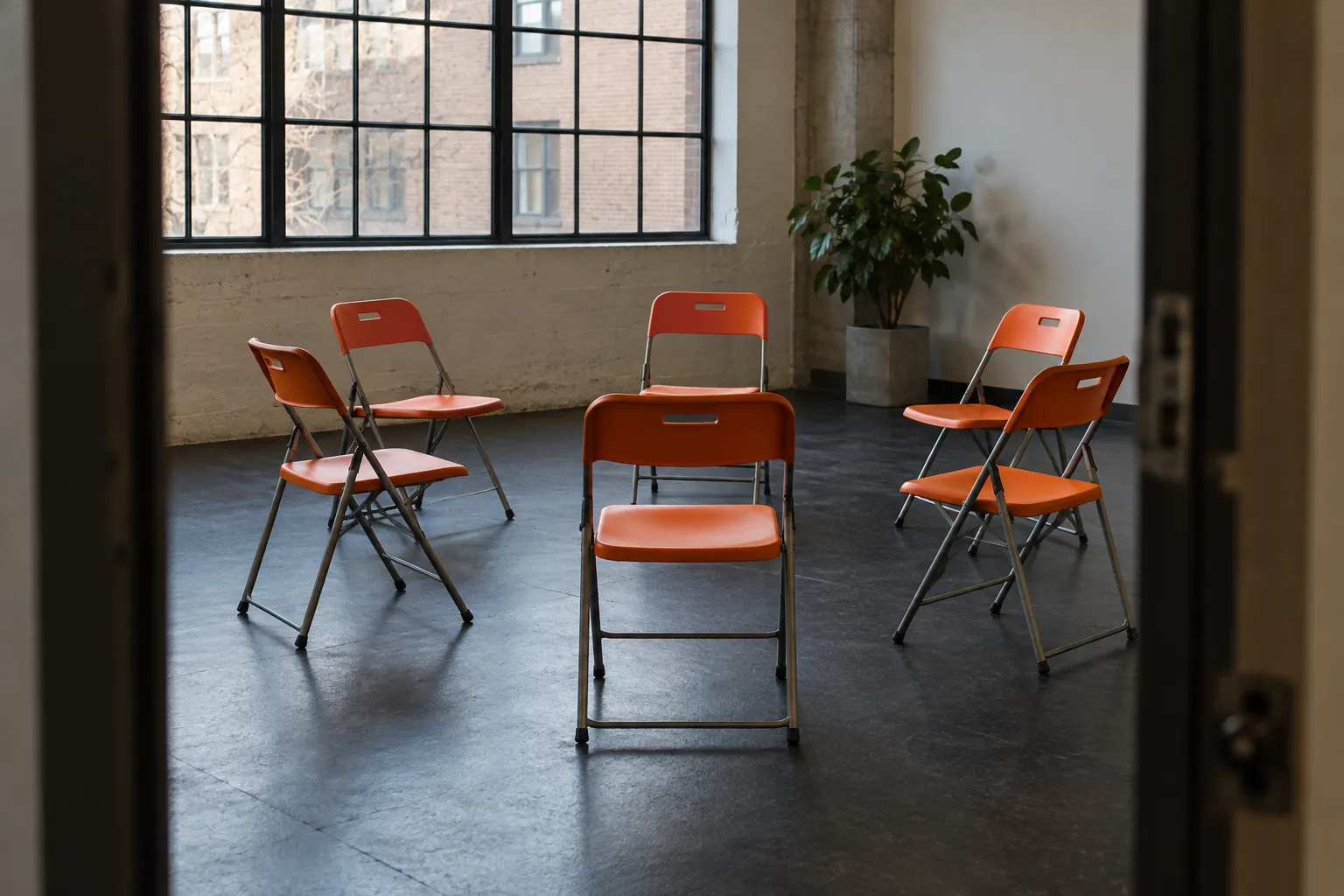Illustration: Orange plastic folding chairs form an imperfect circle on a dark rubber floor beneath an industrial window, photographed from the doorway.