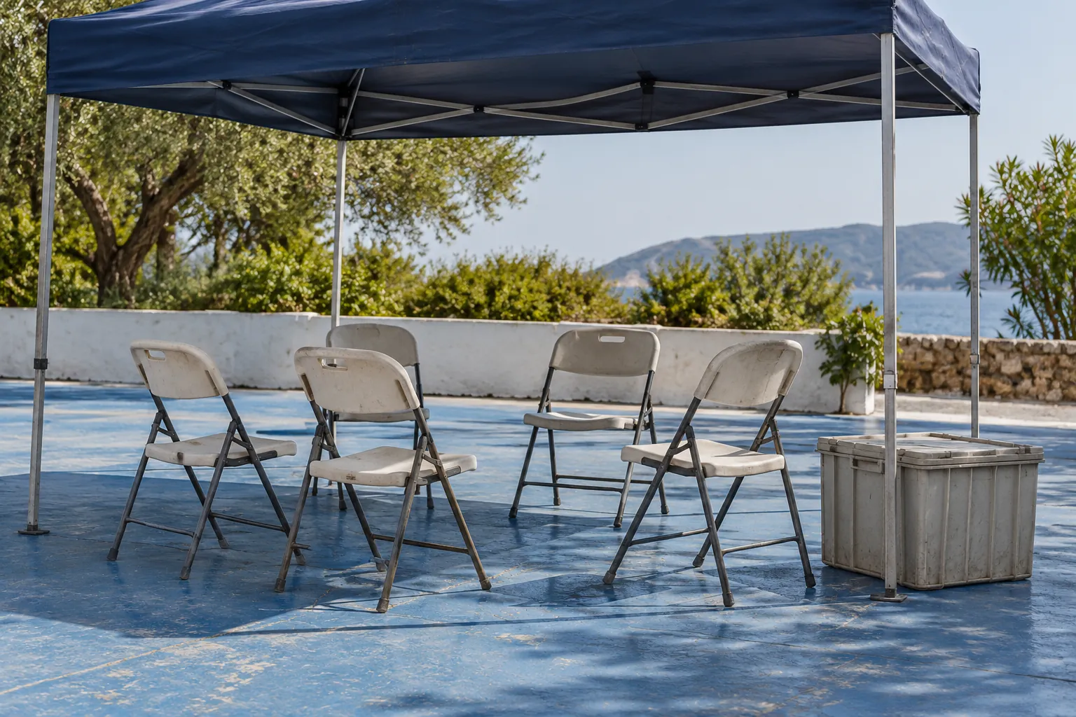 Illustration: A small gathering of folding chairs sits beneath a shade canopy on blue painted concrete, with a plain supply crate, in a wide side view.