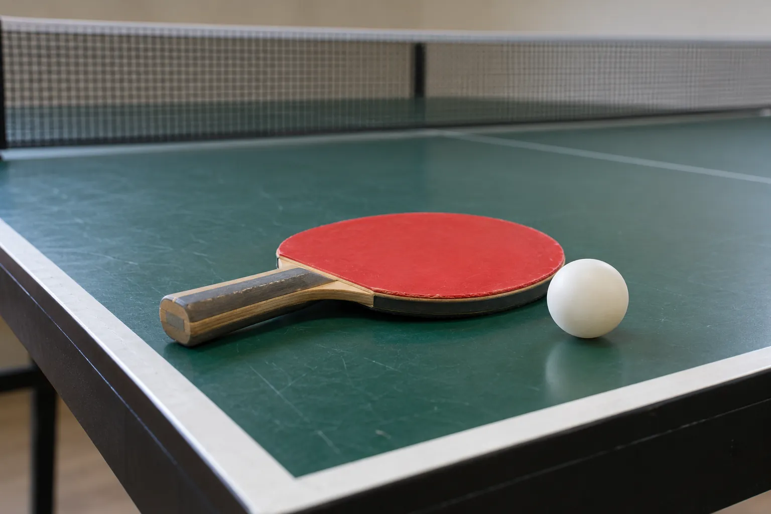 Illustration: A plain table-tennis paddle with red rubber lies beside one white ball on the corner of a green table-tennis table.