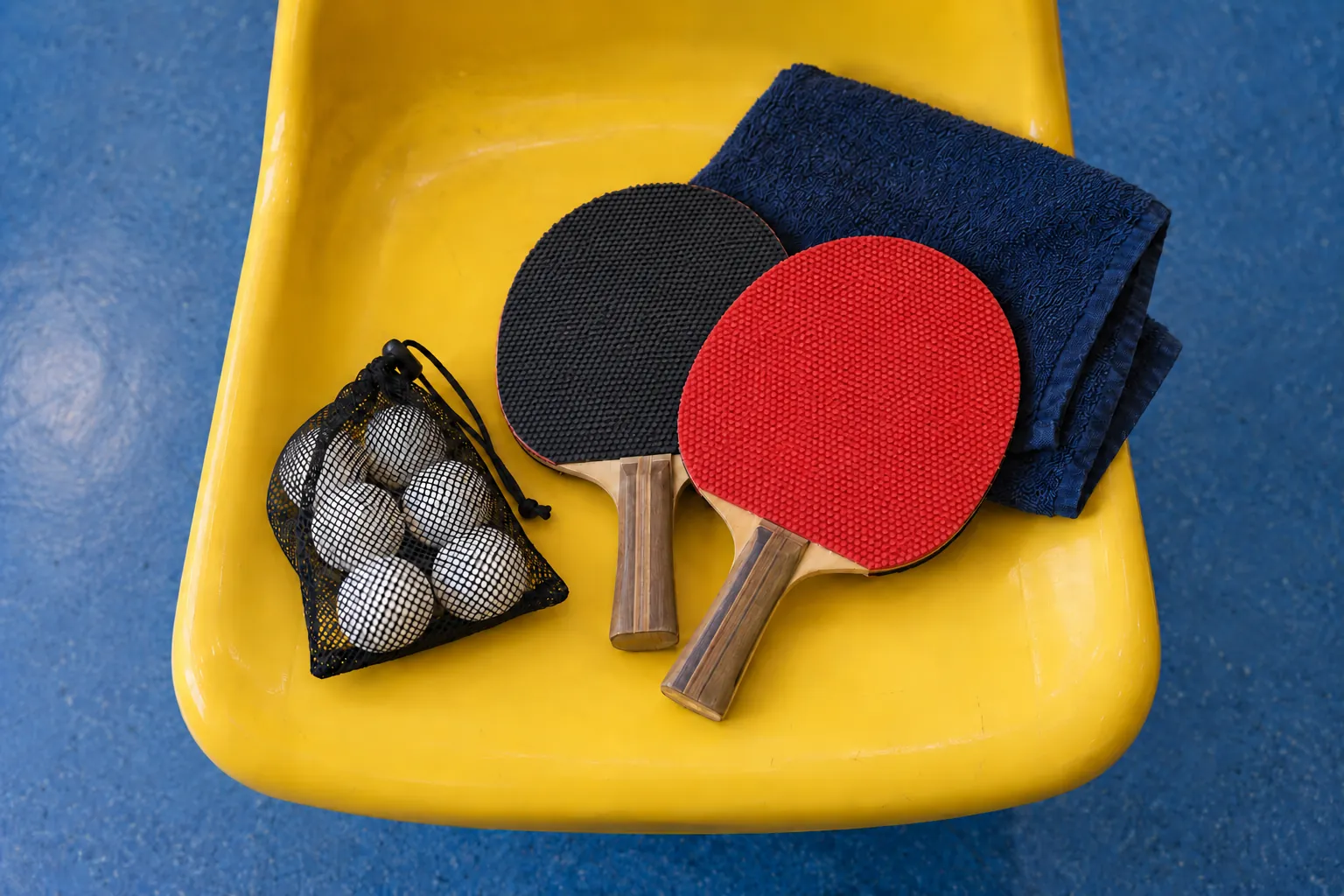 Illustration: Two plain paddle cases, a mesh pouch of white table-tennis balls and a folded navy towel sit on a yellow molded sports seat.
