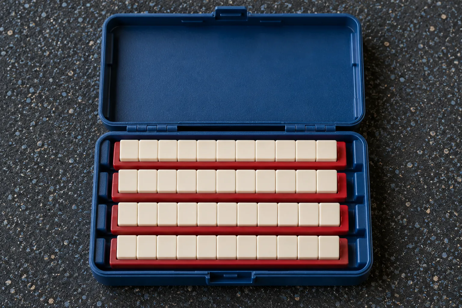 Illustration: Blank ivory game tiles in red racks fit inside an open blue travel case on rubber flooring, shown overhead with physical hinges and realistic scale.