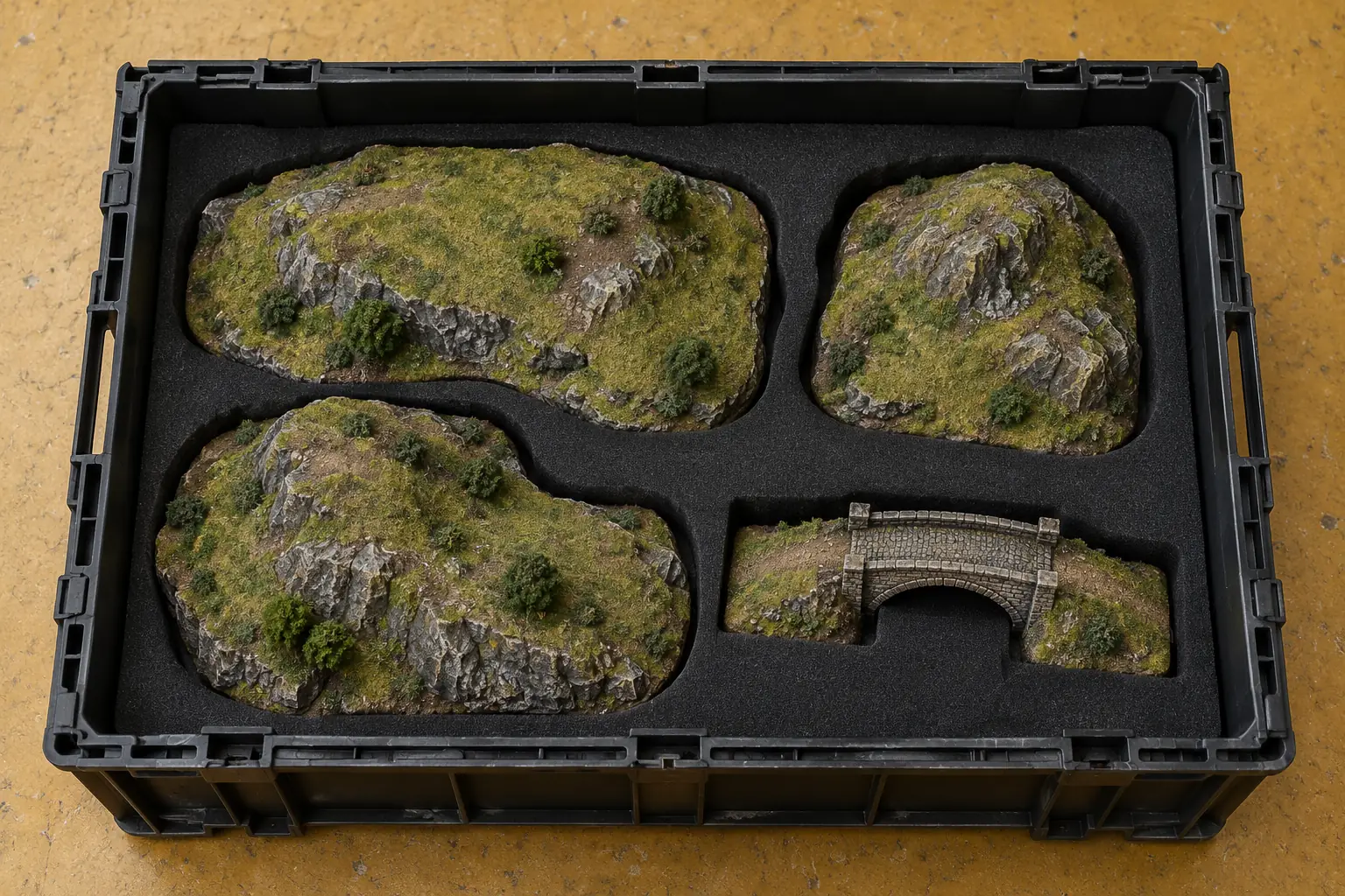 Illustration: Unbranded miniature strategy terrain hills and a simple bridge nest inside a black foam-lined crate on a mustard linoleum floor, photographed overhead.