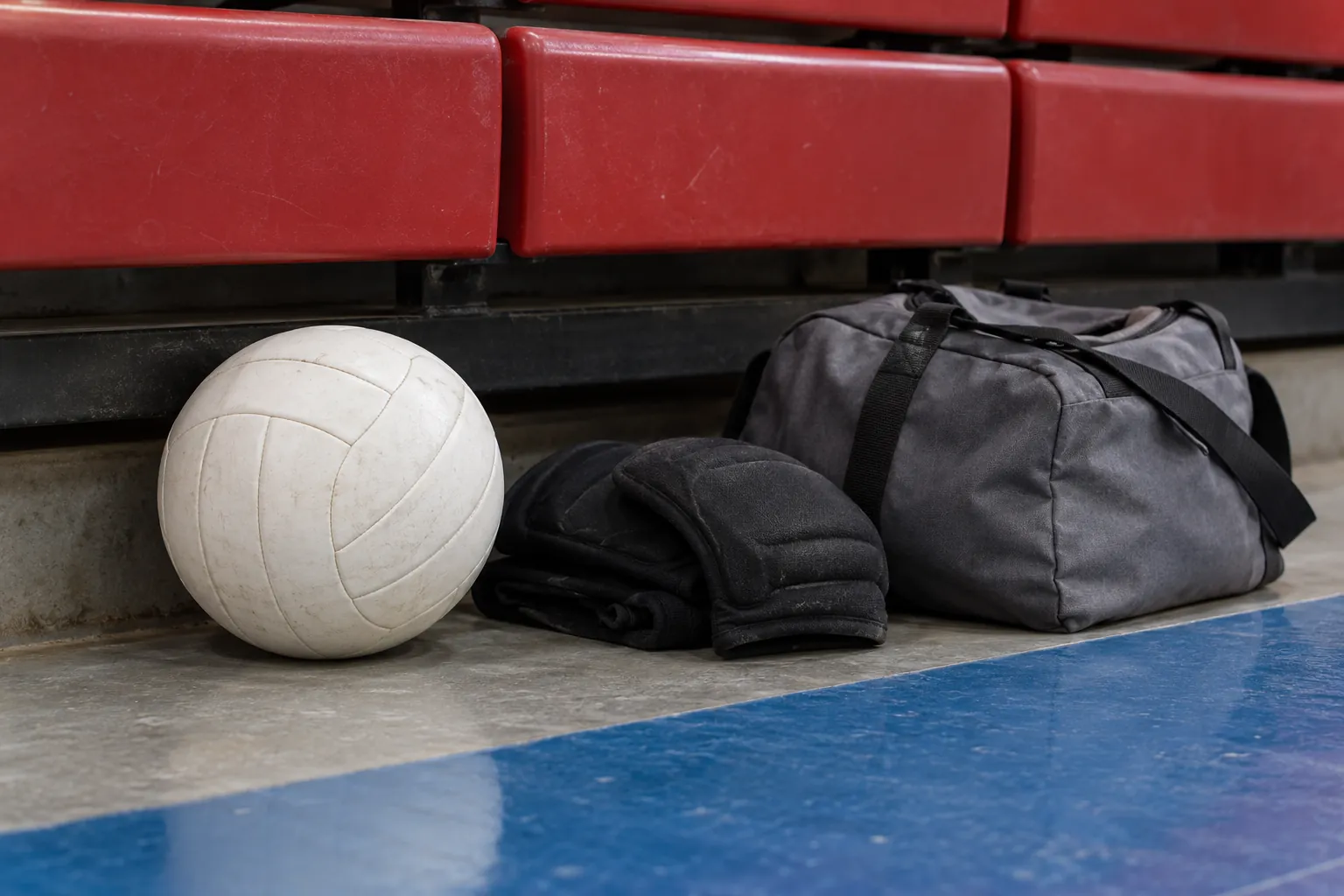 Illustration: A plain volleyball, folded knee pads and a small gray gear bag sit beneath the lowest row of red indoor bleacher seats.