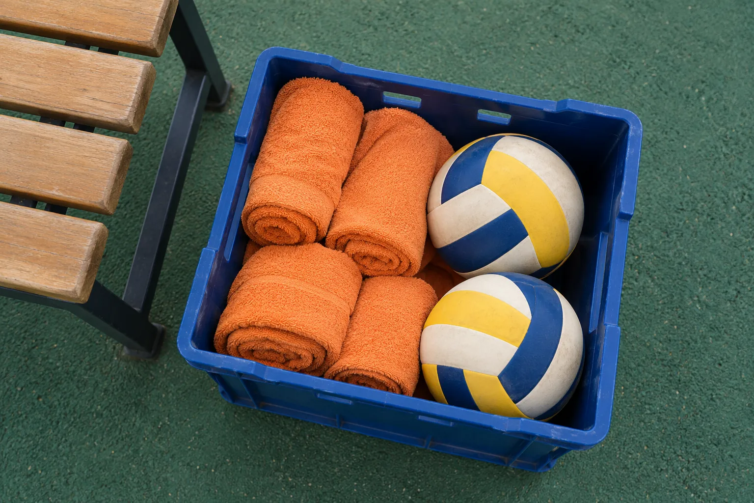 Illustration: Rolled plain orange towels and two unbranded volleyballs fill a blue plastic equipment crate beside a court-side bench.