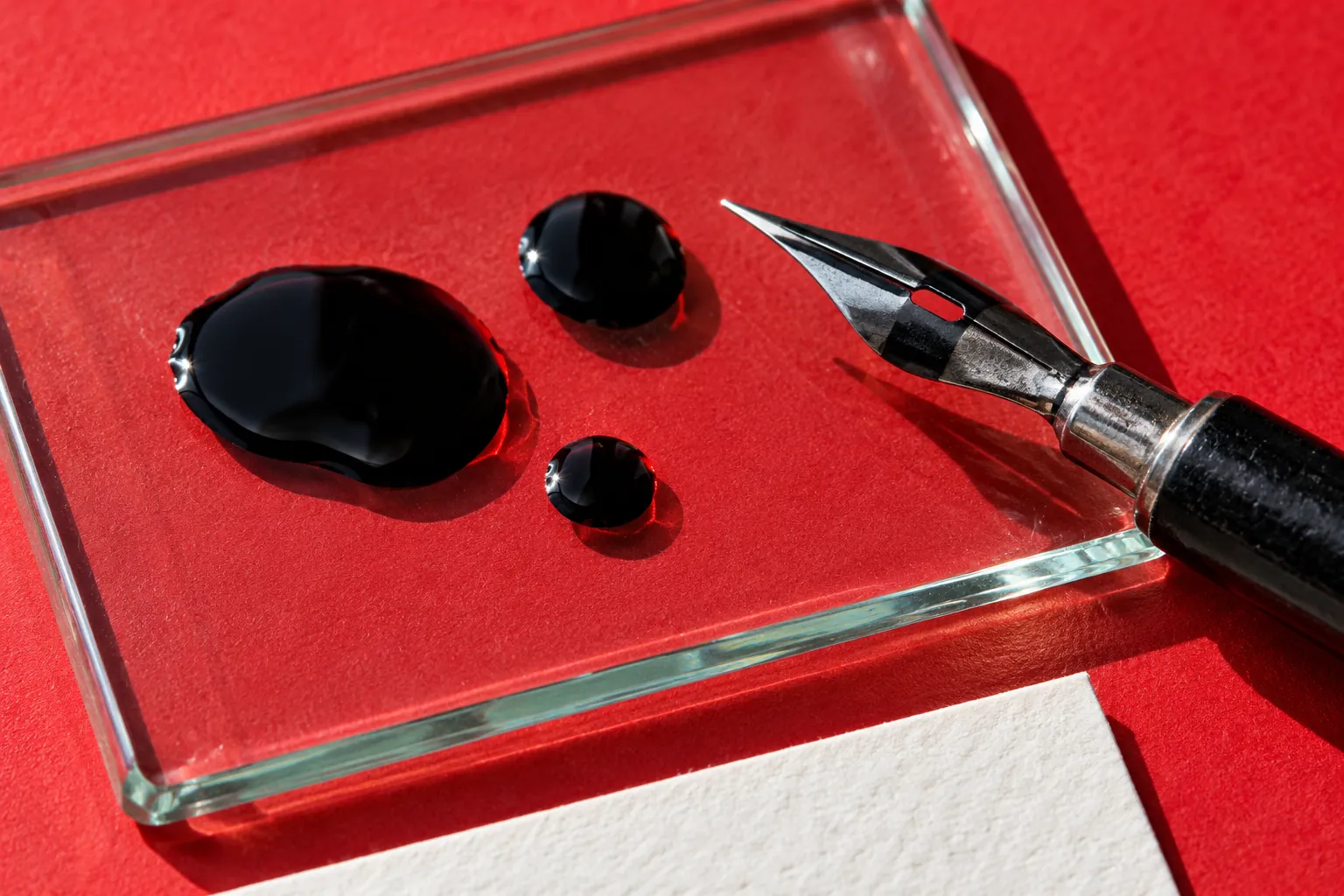 Illustration: Black ink drops and a dip-pen nib on glass above red paper.